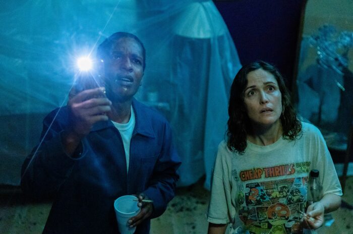 A film still of a nighttime scene of a Black man holding a cup in one hand and a holding up a phone with flashlight on in the other, standing next to a white woman with wet hair and a Cheap Thrills t-shirt on.