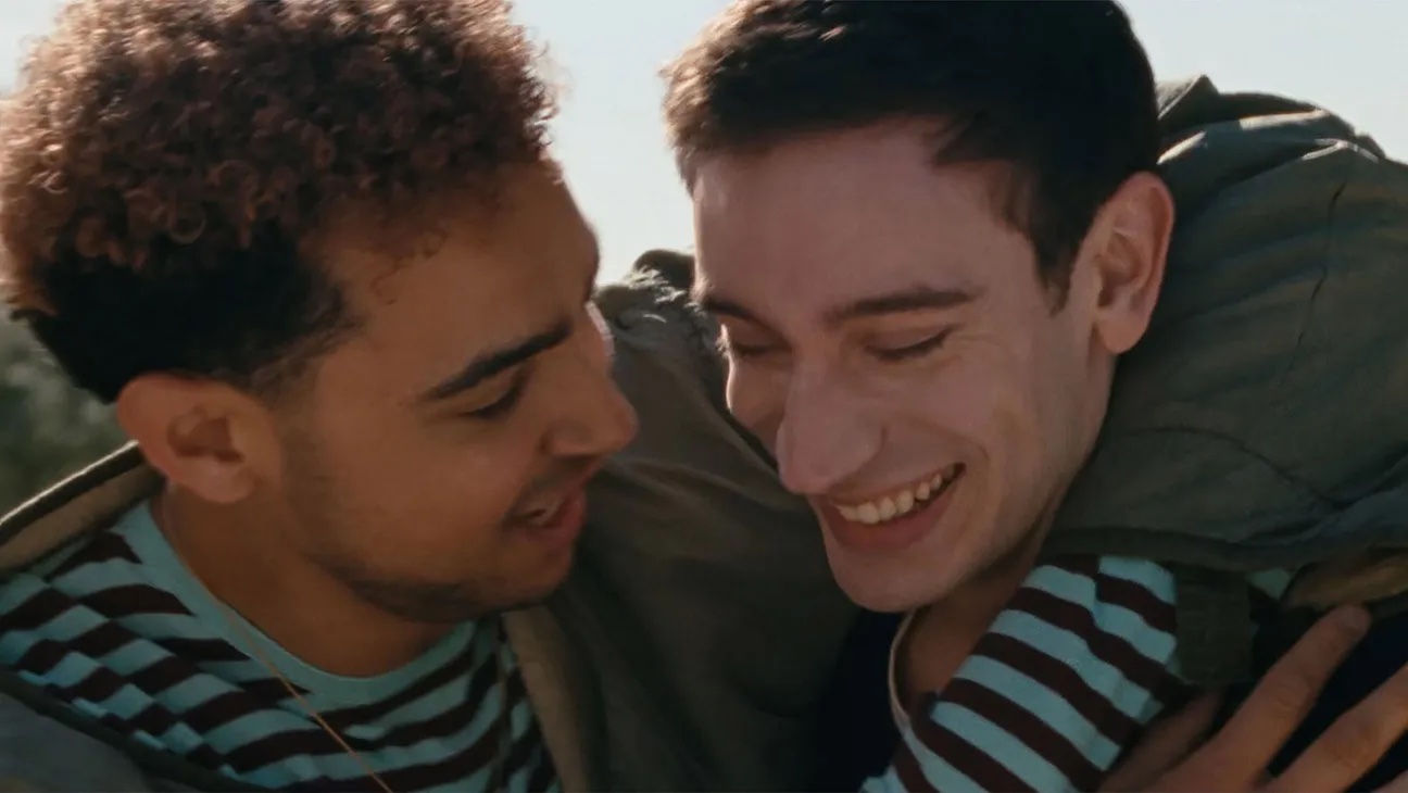 A film still of two young men smiling. One has his arm around the other's neck.