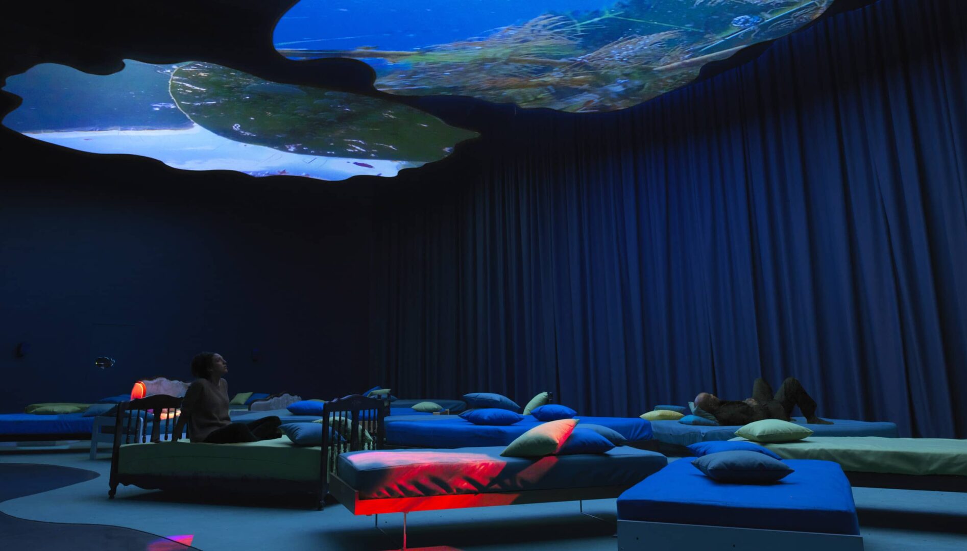 Two people reclining on beds in a gallery space, viewing a projection on the ceiling.