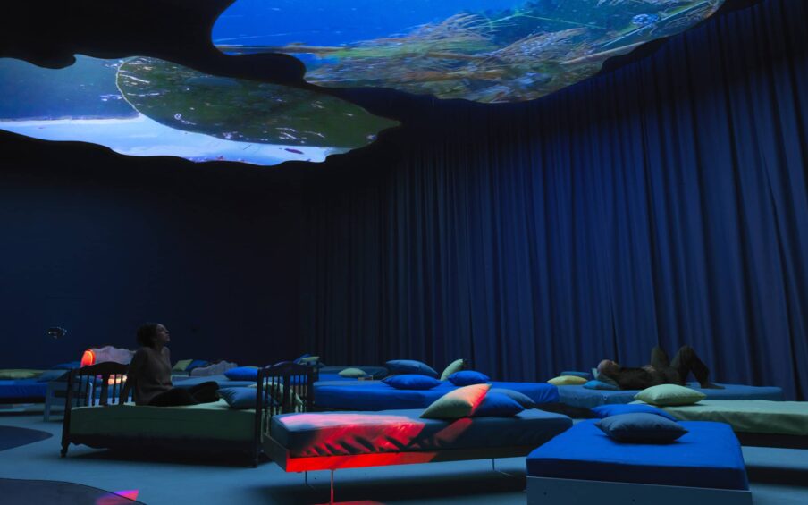 Two people reclining on beds in a gallery space, viewing a projection on the ceiling.