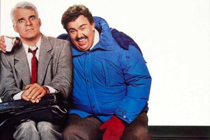 Film still of two white men sitting on a bench. On the left, a man with white hair wearing a grey suit (Steve Martin) has a scared look on his face. On the right is a man with a mustache and a blue jacket, smiling with his arm around the other man.