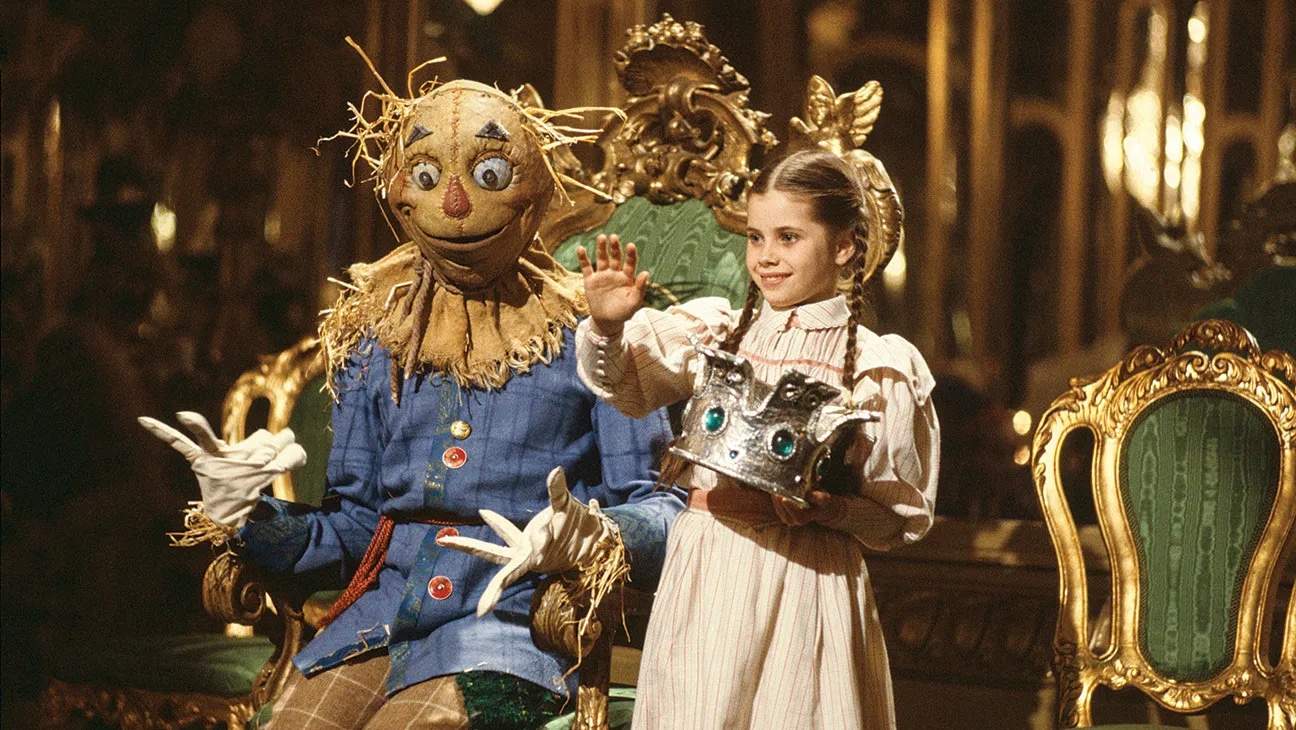 Film still of a scarecrow sitting on a throne and a young girl with long brown braids holding a crown.