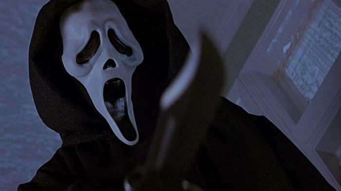Film still of a person in a black hooded gown and a white scream mask, holding a knife.