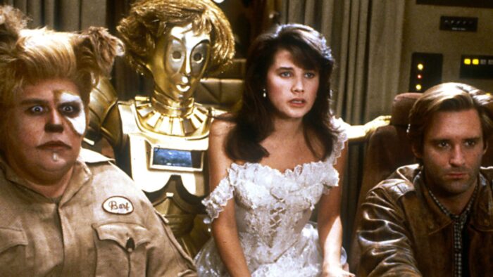 Film still from the movie Spaceballs. A man in a dog costume, a gold robot, a woman with long brown hair and a white dress, and a man in a leather jacket sit in the cockpit of a spaceship.