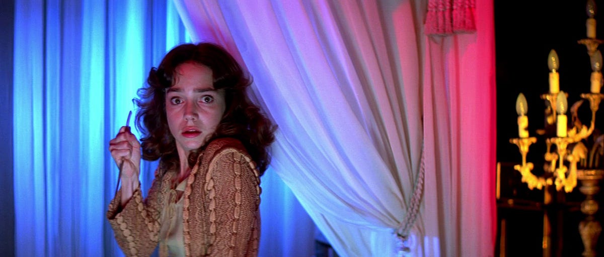 Film still of a white woman with long curly brown hair in front of a white curtain lit with blue and red lights. She has a scared look on her face and is holding a knife up.