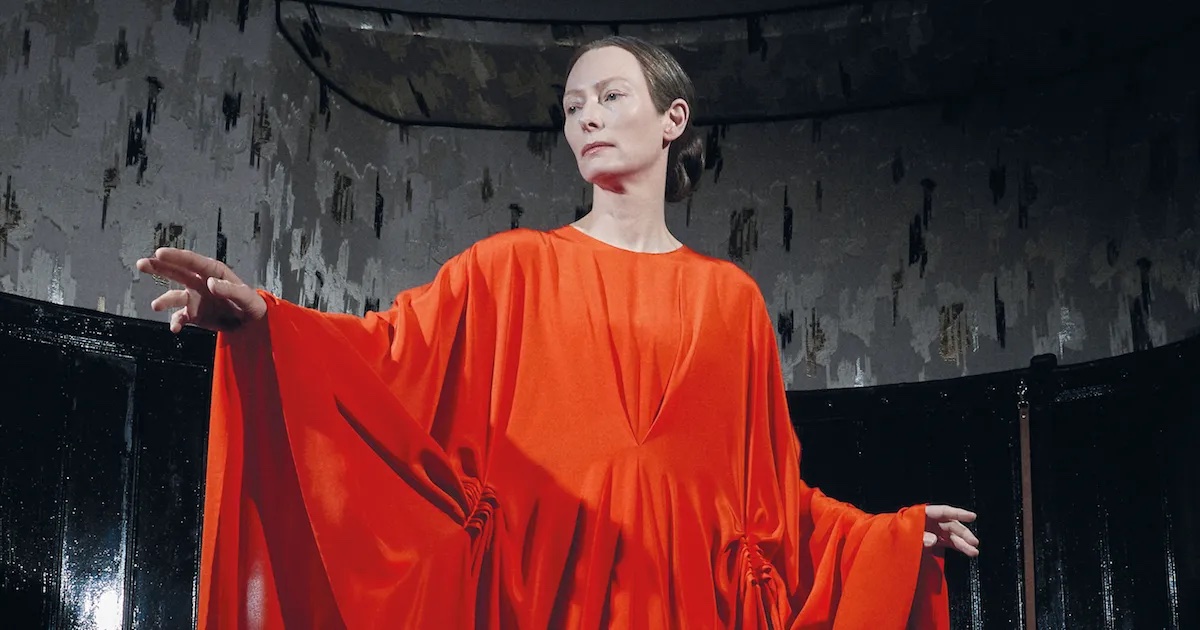 Film still of a white woman (Tilda Swinton) with her hair pulled back, wearing a long, draping orange dress and holding her arms out.