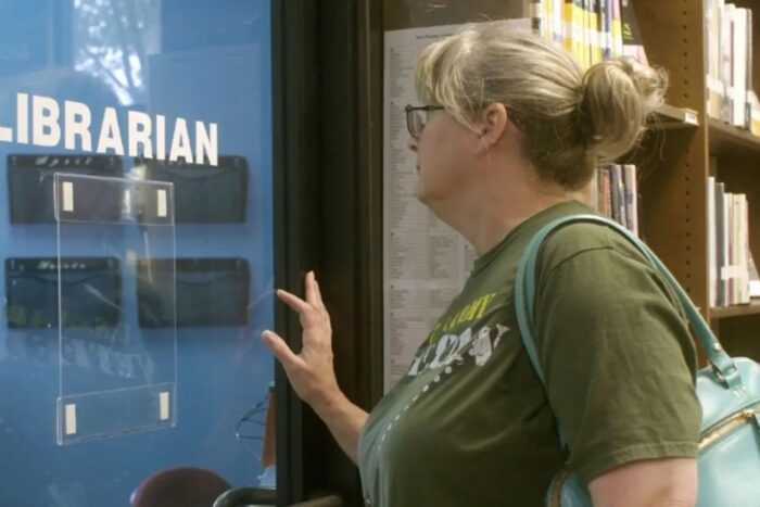 A white woman with long blond hair in a bun, wearing eyeglasses and a green t-shirt, pushing on a door that says "Librarian."