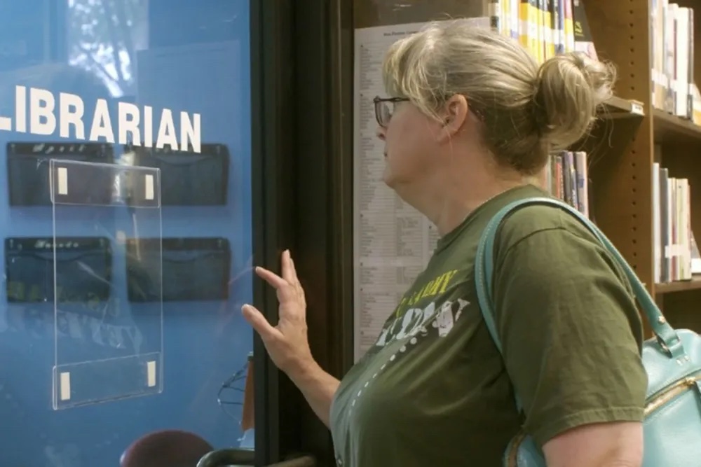 A white woman with long blond hair in a bun, wearing eyeglasses and a green t-shirt, pushing on a door that says "Librarian."