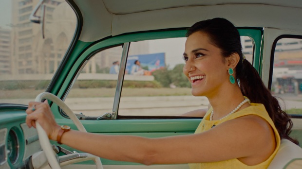 A smiling young Indian woman with long brown hair in a ponytail, turquoise earrings and a pearl necklace, driving a seafoam green car.