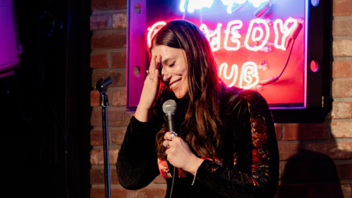 A white woman with long brown hair on stage at a comedy club. She is smiling, has her eyes closed, and is holding one hand up to her head in seeming embarassment.