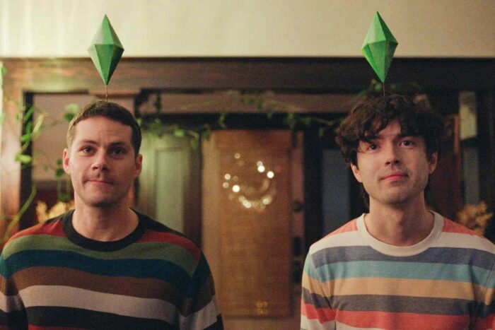 Film still of two young white men, both in multicolored striped sweaters, each wearing a green diamond headpiece.