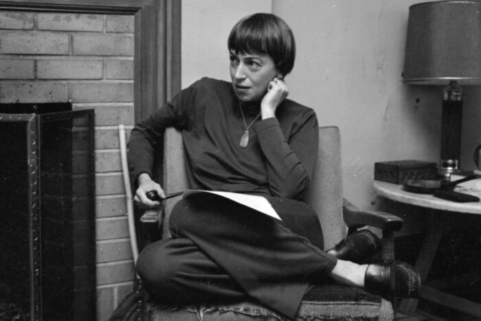 A black and white photo of the author Ursula K LeGuin sitting on a chair with her legs curled up and papers on her lap.