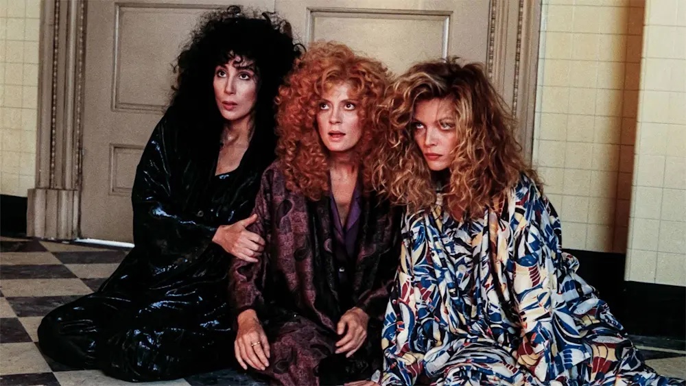 Three white women (Cher, Susan Sarandon, and Michelle Pfeiffer), all with long curly hair, sitting on a black and white checkered floor, with tired or scared looks on their faces.