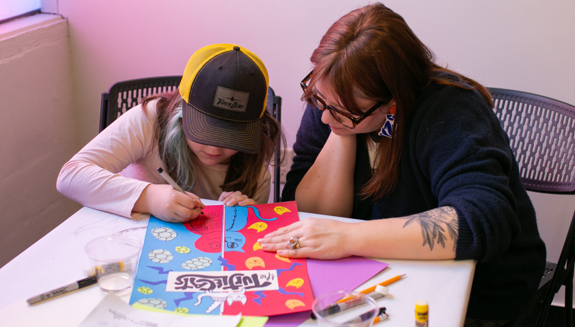 An adult woman with eyeglasses and long hair leaning over a table with a young person while long blue hair and a baseball cap works on a poster. There are craft supplies scattered on the table.