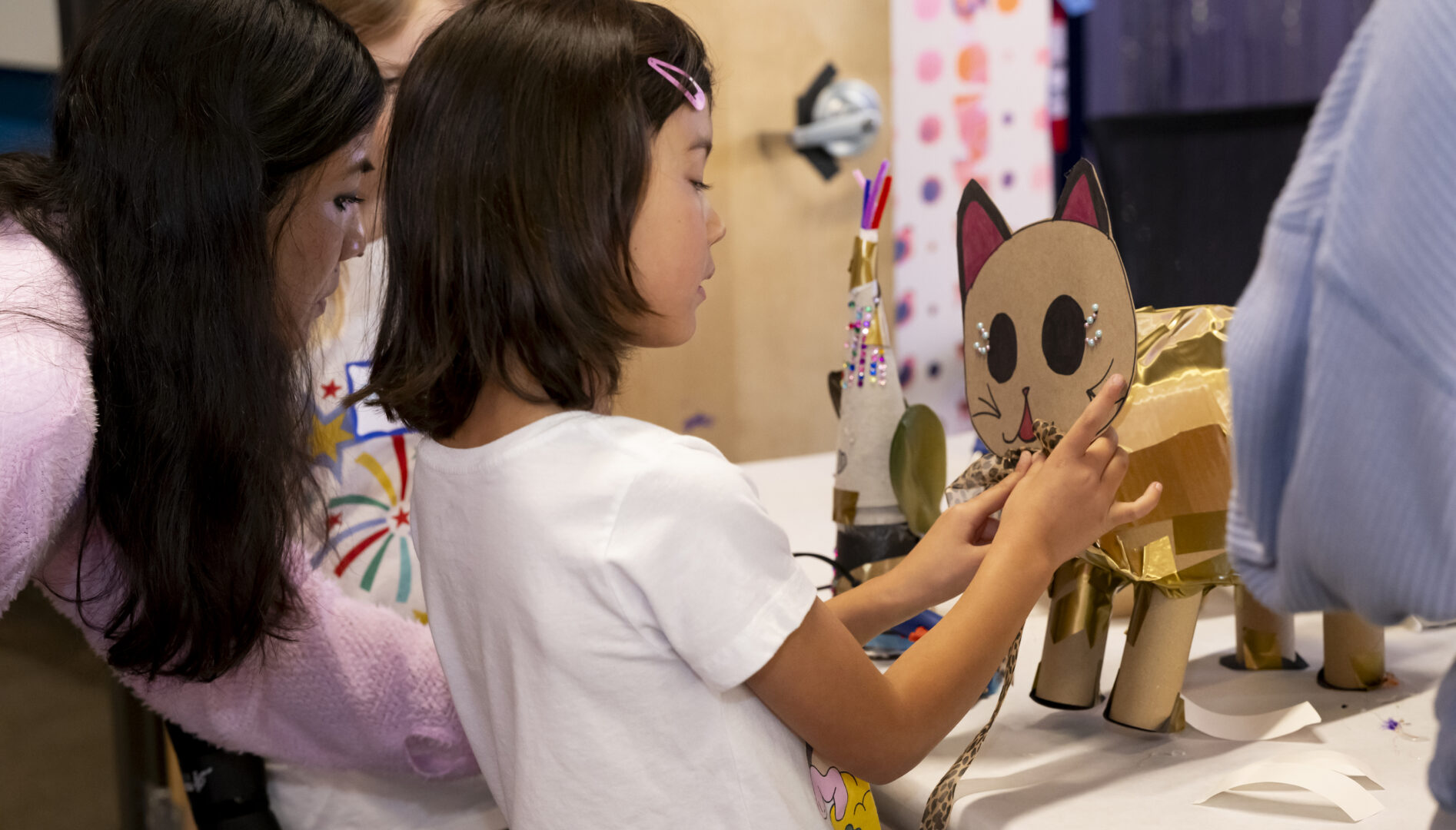 A young girl with dark brown hair works on a cardboard cat that she's crafting.