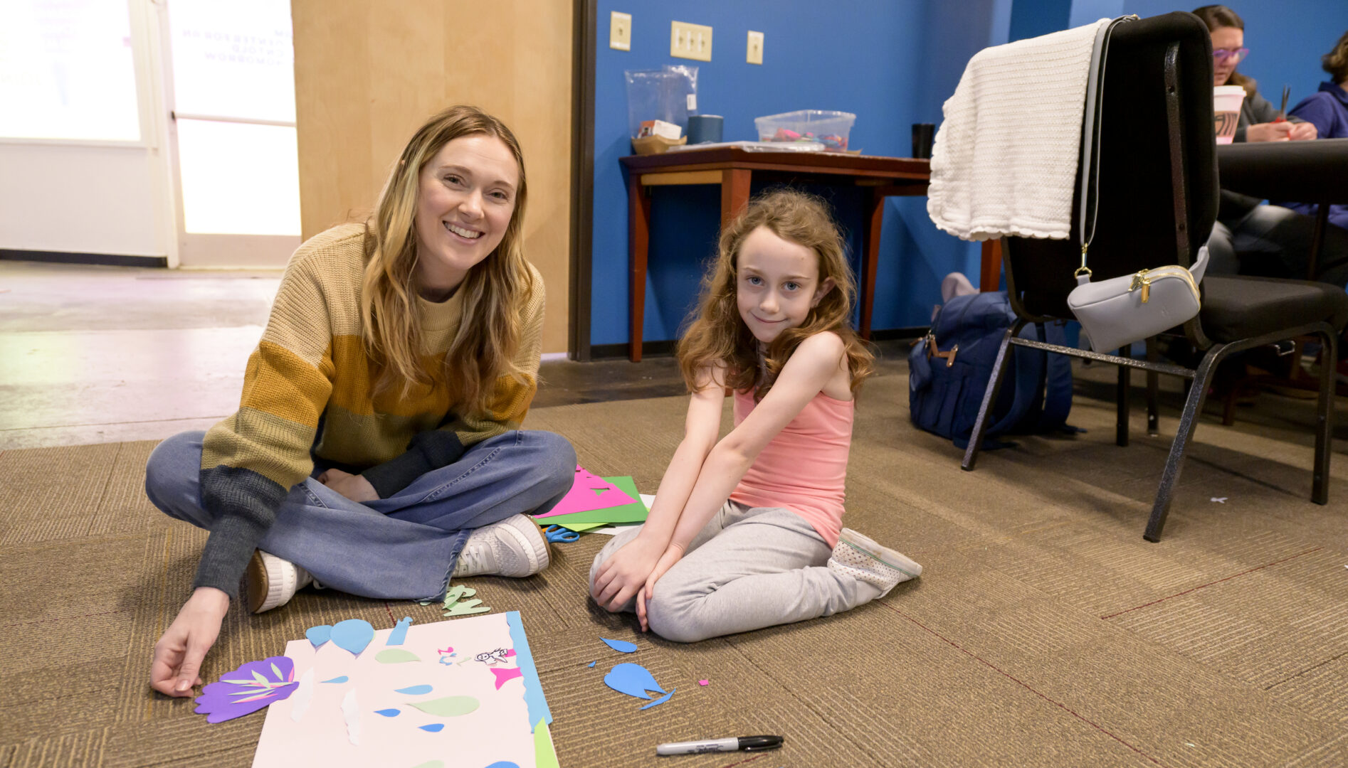 An adult woman with long blond hair and a young girl with long red hair sitting on the floor, smiling, with paper and markers on in front of them.