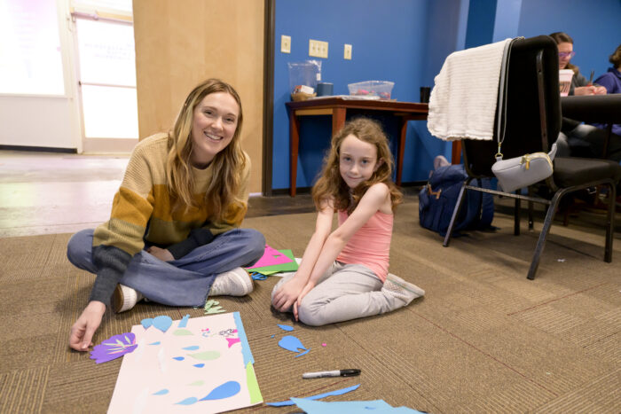 An adult woman with long blond hair and a young girl with long red hair sitting on the floor, smiling, with paper and markers on in front of them.