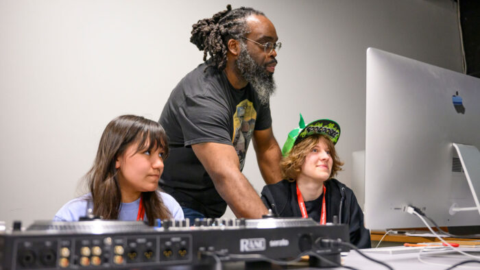 A man with dreadlocks and eyeglasses working with two kids at a computer and DJ mixing table.