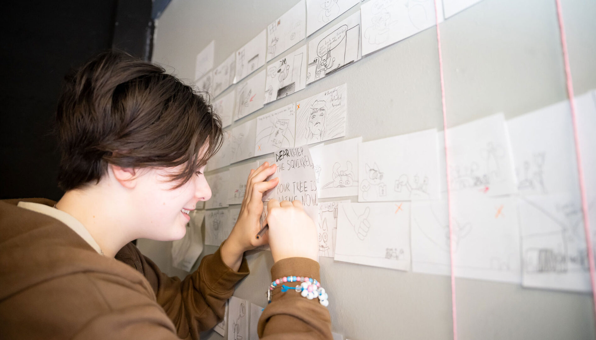 A kid wearing a brown sweatshirt drawing on paper pinned up on a whiteboard.