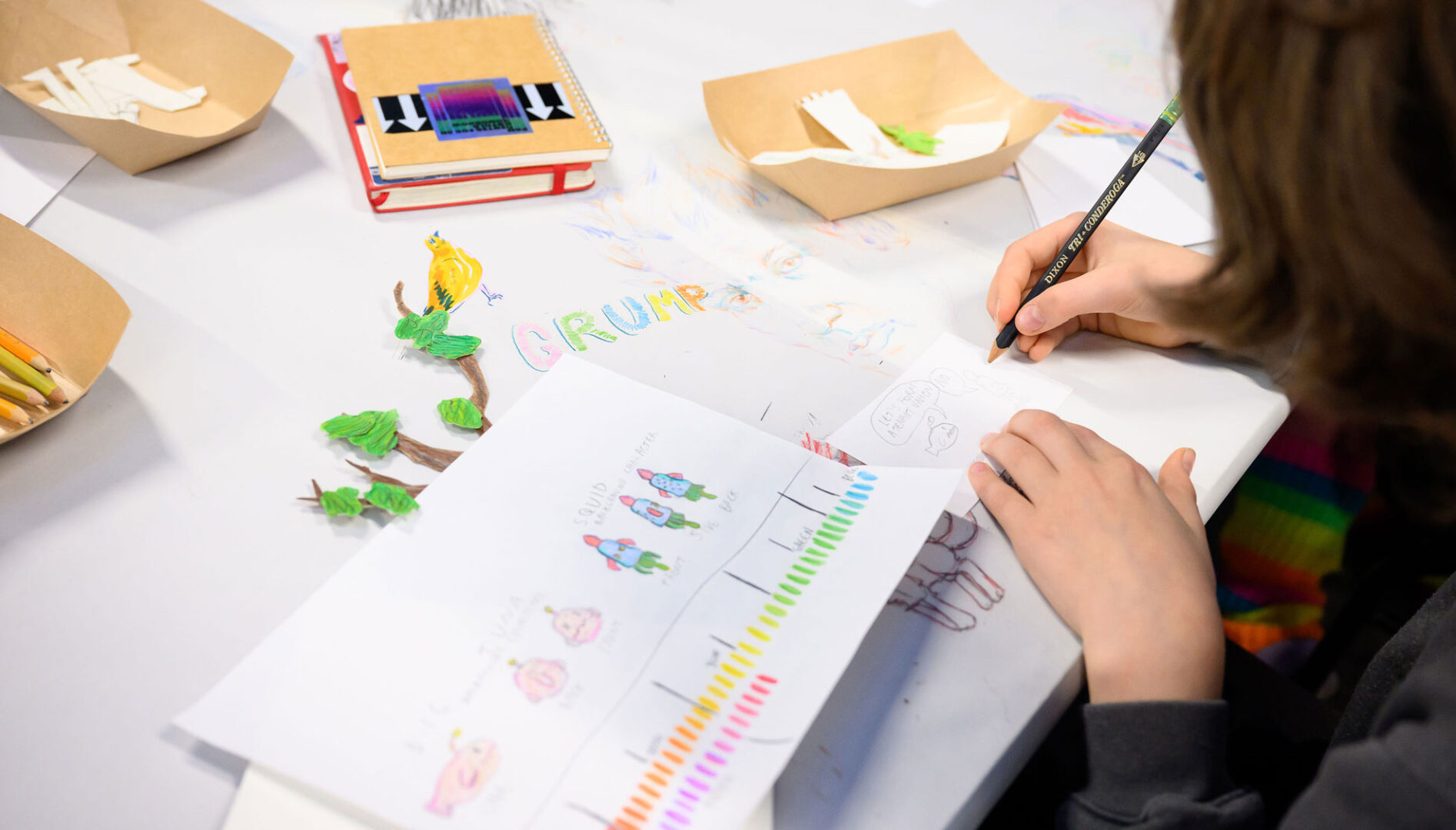 Photo of a kid drawing at a table of paper, colored pencils, and art supplies.