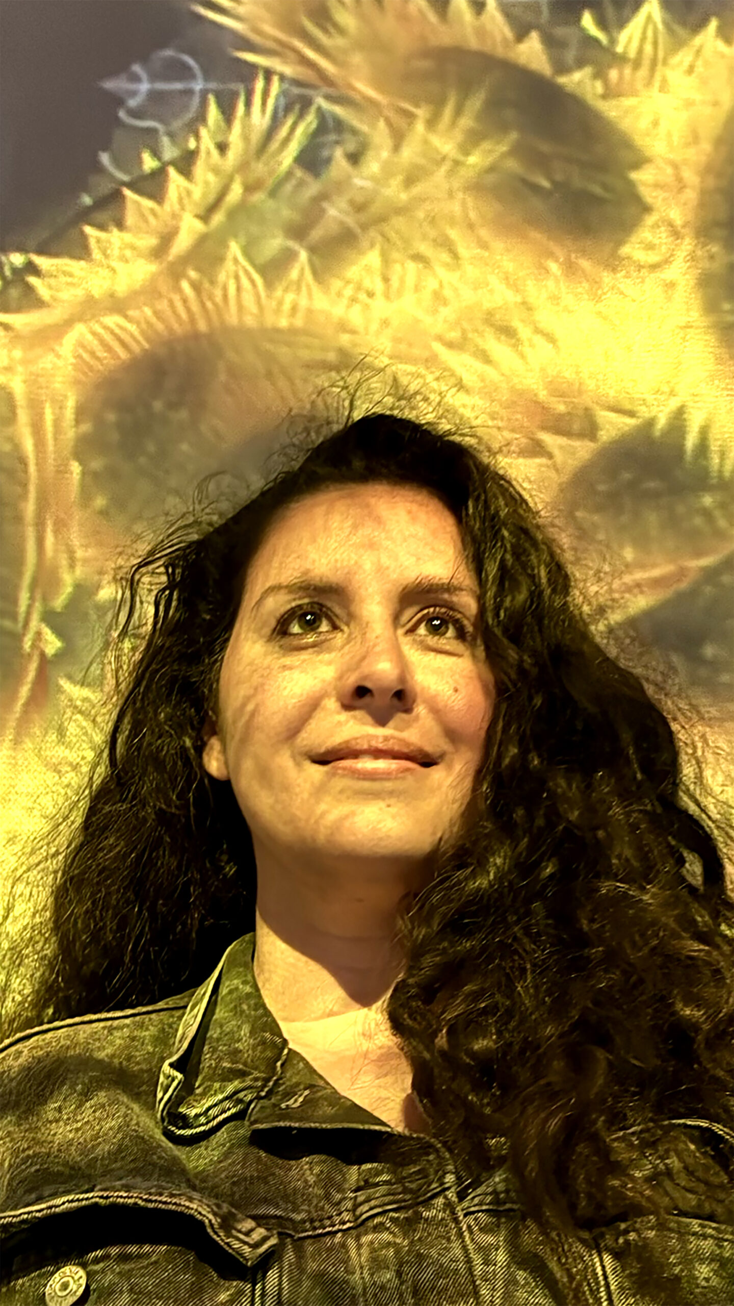 Portrait of a smiling woman with long dark, wavy hear against a projection of sunflowers.