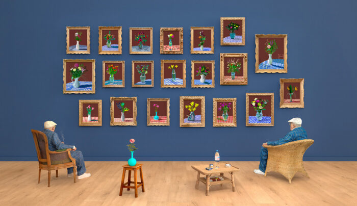 A blue gallery wall with a grid of framed art on it. Looking towards the wall are two chairs and two tables. David Hockney sits in the lefthand chair.