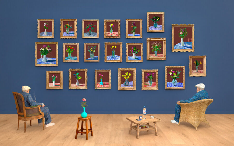 A blue gallery wall with a grid of framed art on it. Looking towards the wall are two chairs and two tables. David Hockney sits in the lefthand chair.