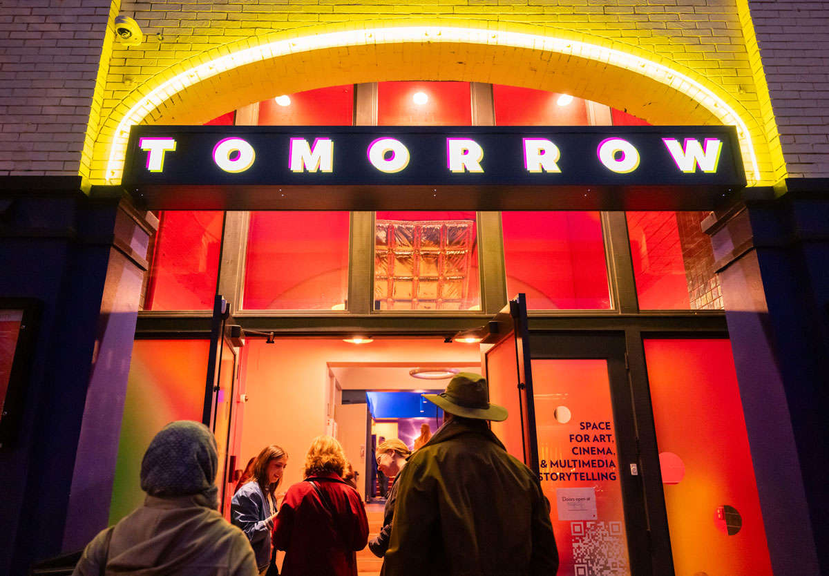 Photo of the outside of the Tomorrow Theater at night with people walking in through the front door.