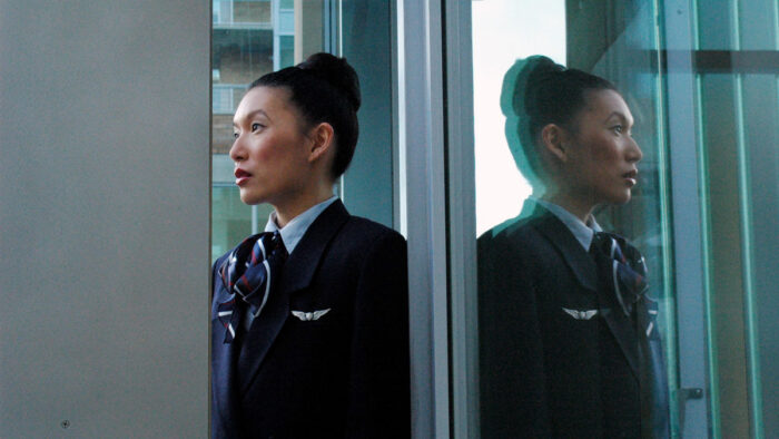 A film still of a woman in a uniform with her hair in a bun. She is standing in front of and reflected in a large pane of glass.