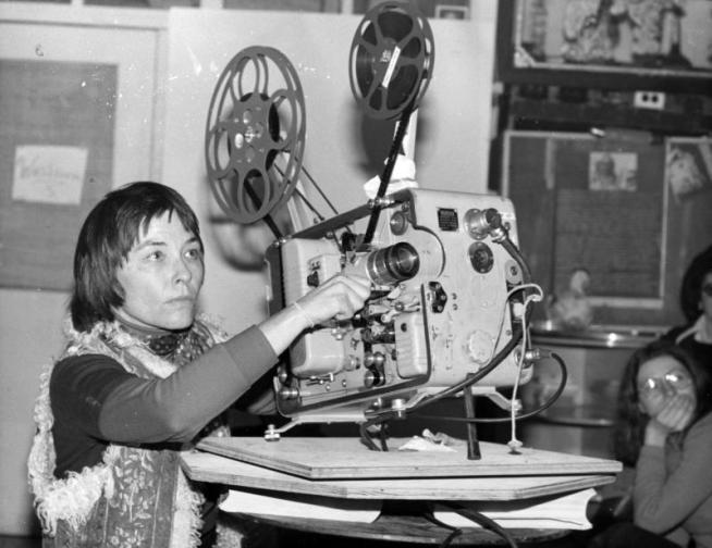 Black and white photo of a woman standing next to a reel-to-reel film projector.