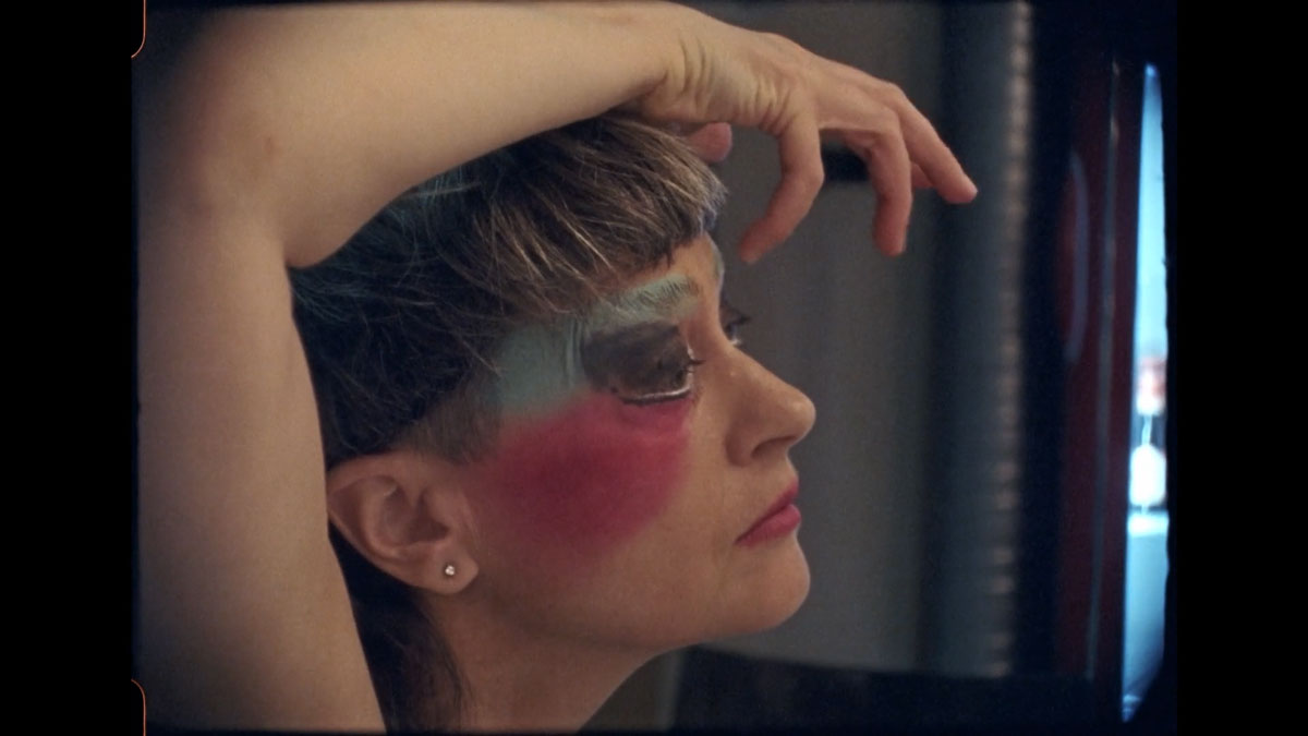 Film still of a woman viewed from the side. She is wearing blue and pink makeup around her eye and cheeks and is arching her arm up around her head to touch her face.