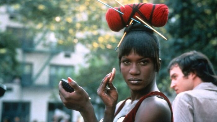 Film still of a Black drag queen with a headpiece that looks like yarn and knitting needles. She is holding up a compact and a powder applicator to her face and looking towards the camera.
