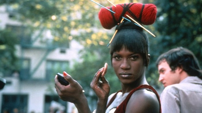 Film still of a Black drag queen with a headpiece that looks like yarn and knitting needles. She is holding up a compact and a powder applicator to her face and looking towards the camera.