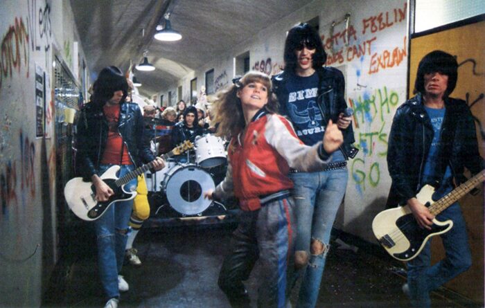 Film still of the Ramones playing in a high school hallway. A young woman is dancing in front of them.