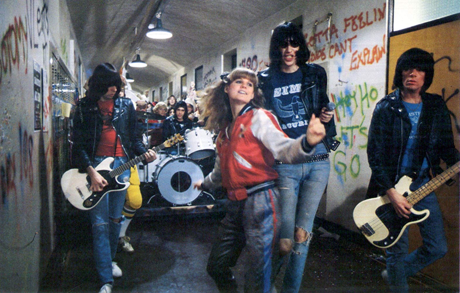 Film still of the Ramones playing in a high school hallway. A young woman is dancing in front of them.