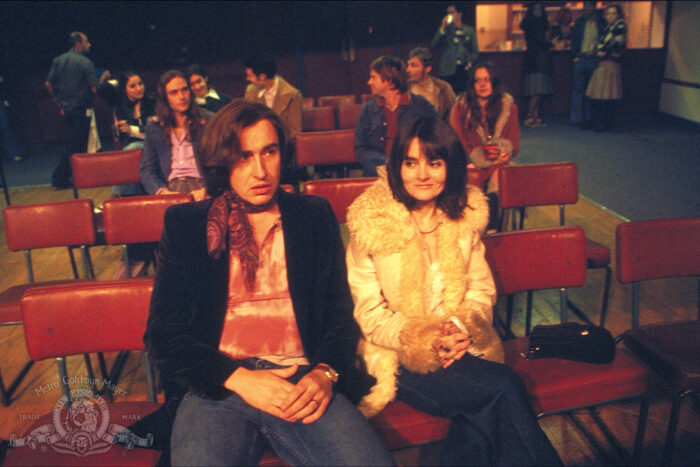 Film still of two people in 1970s outfits sitting in red chairs.