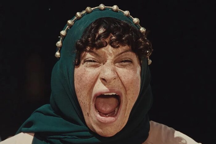 Close up of a woman yelling with her mouth wide open. She is wearing a green headscarf.