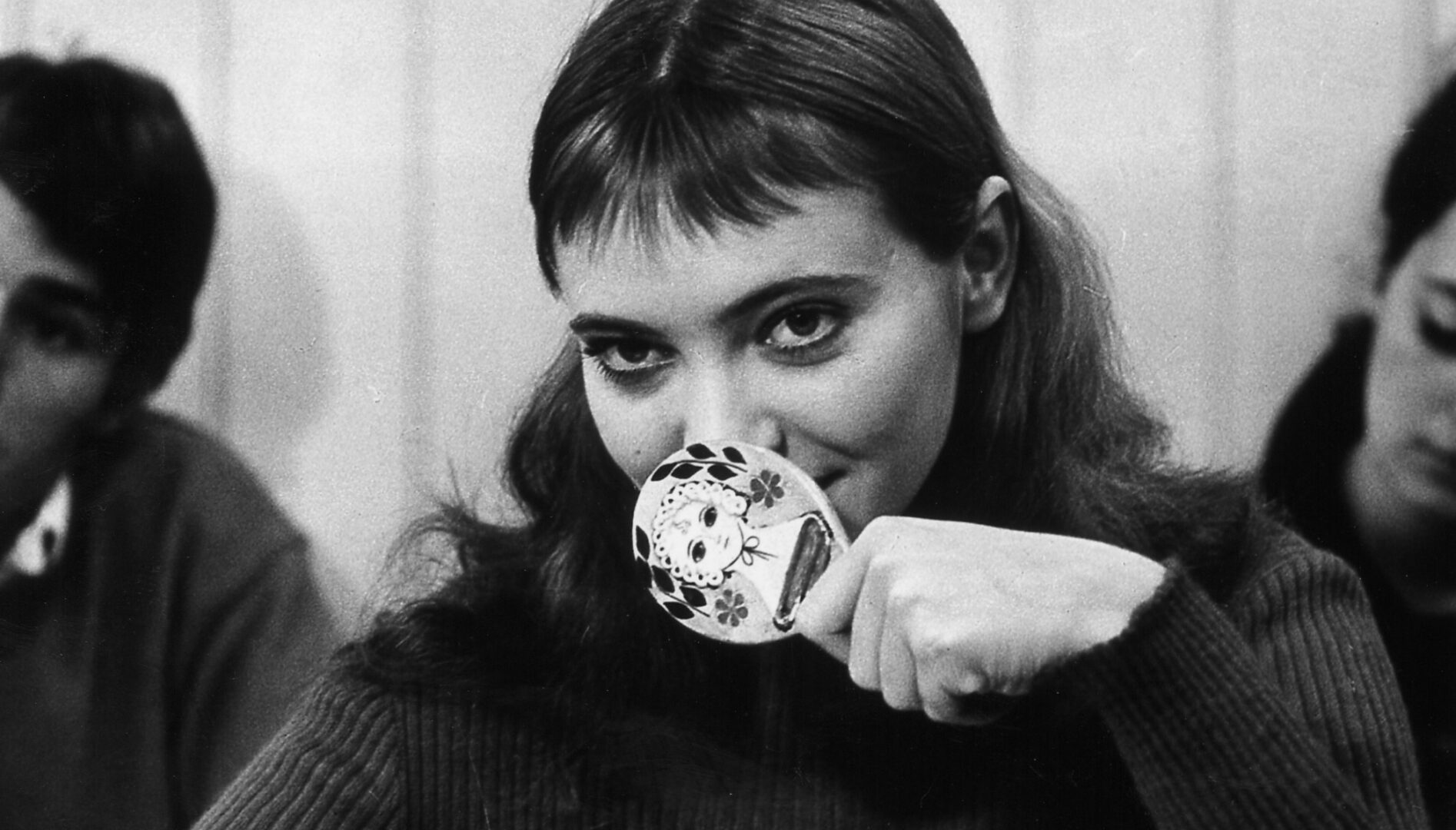 A black and white film still of Anna Karina holding a small mirror up in front of her mouth.