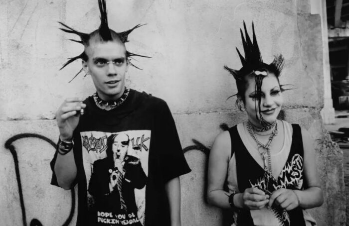 Black and white film still of a two punk rockers with spiky hair against a graffitied wall.