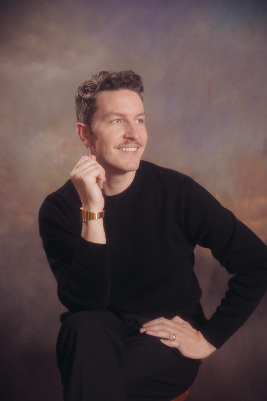 Studio photo of a man with brown hair and a mustache, wearing a black sweater and a gold watch.