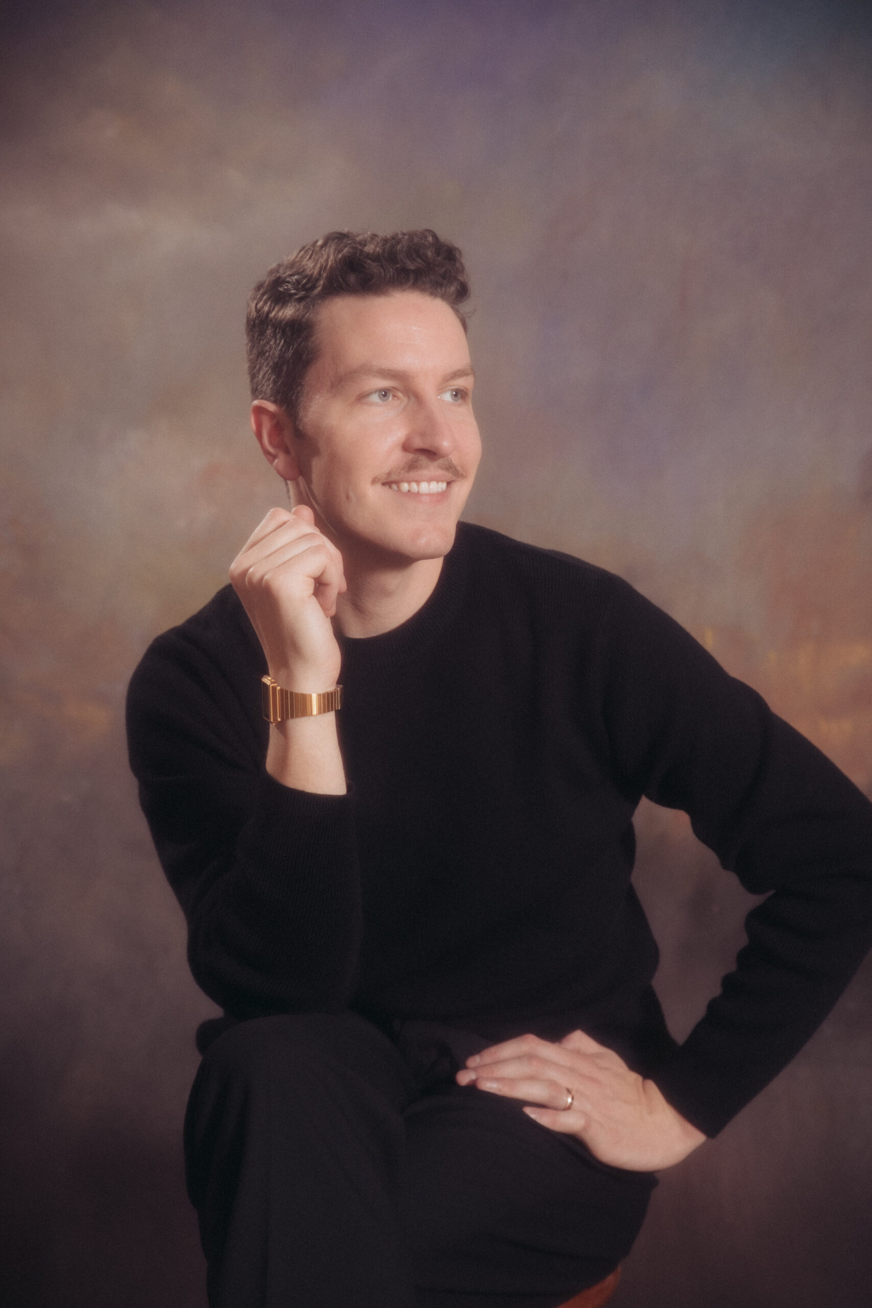 Studio photo of a man with brown hair and a mustache, wearing a black sweater and a gold watch.