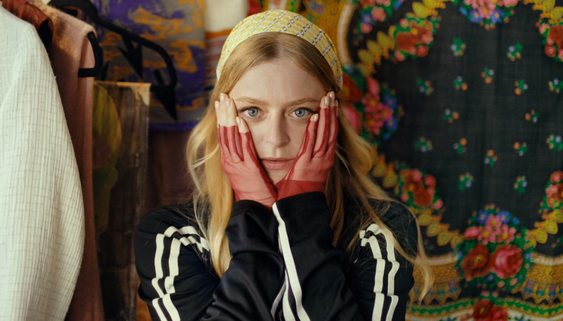 A woman with long blonde hair, wearing a black track jacket with white stripes on the sleeves, holding her hands up to either side of her face.