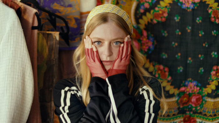 A woman with long blonde hair, wearing a black track jacket with white stripes on the sleeves, holding her hands up to either side of her face.
