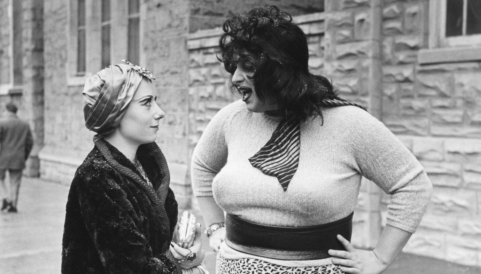 Black and white film still with Divine and a woman with a head scarf talking in the street.