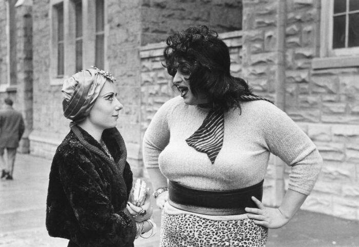 Black and white film still with Divine and a woman with a head scarf talking in the street.