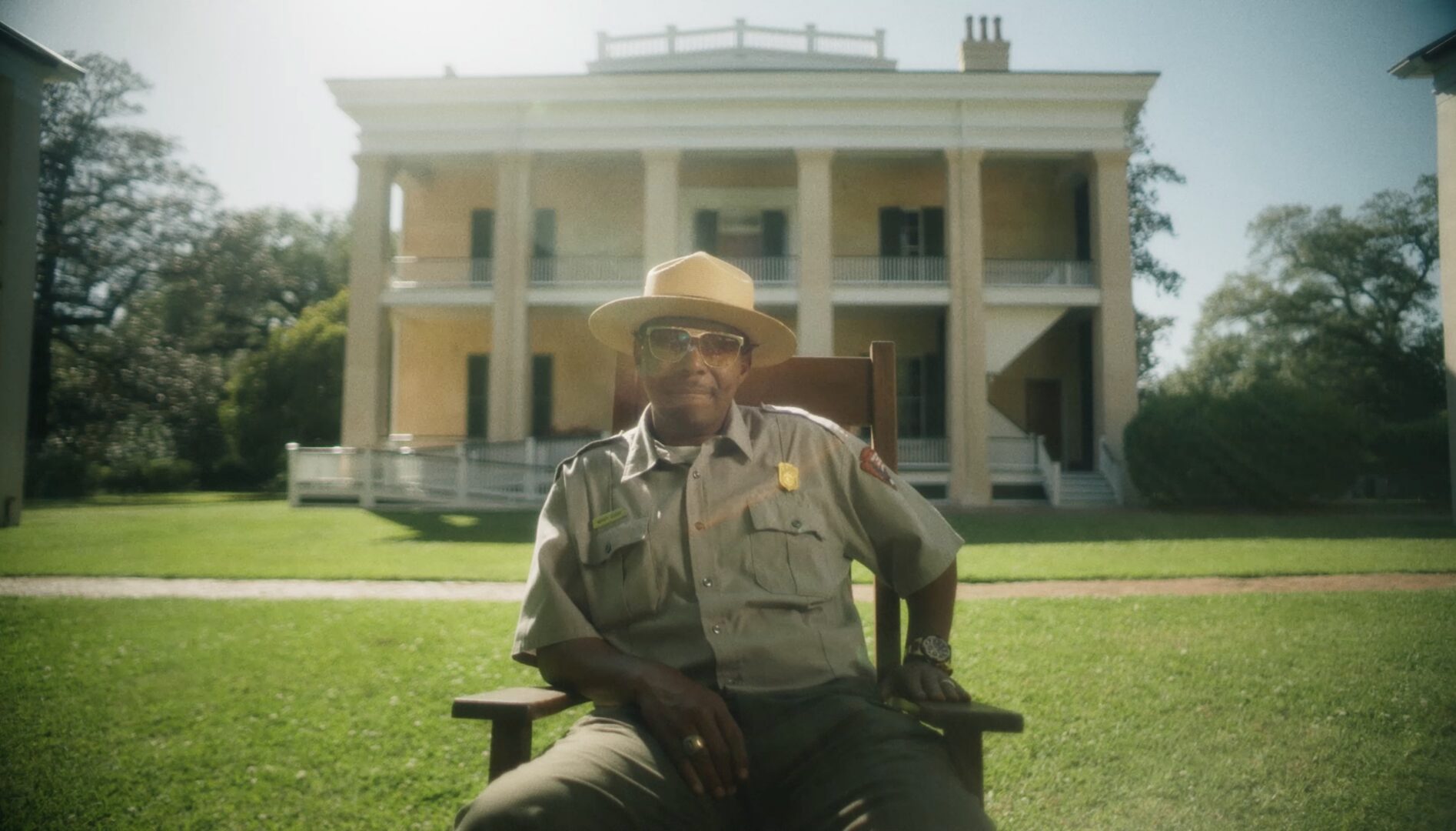 A Black park ranger wearing sunglasses and sitting in a chair in front of an old mansion.
