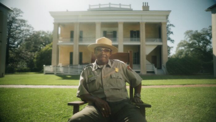 A Black park ranger wearing sunglasses and sitting in a chair in front of an old mansion.