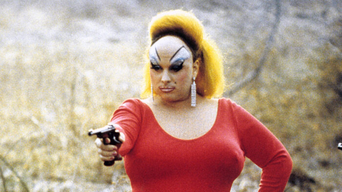Film still of Divine in a red dress, a yellow wig, and dramatic eye makeup. She's holding a gun.