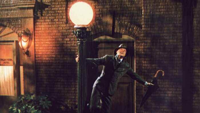 A man in a suit and hat, holding an umbrella and dancing in the rain around a light pole.