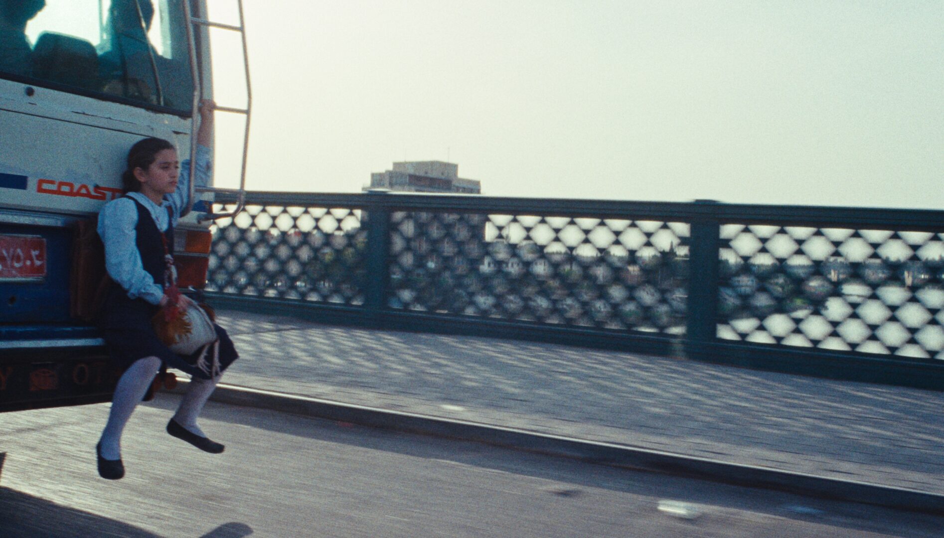 Film still of a little girl sitting on the back of a bus, going over a bridge.